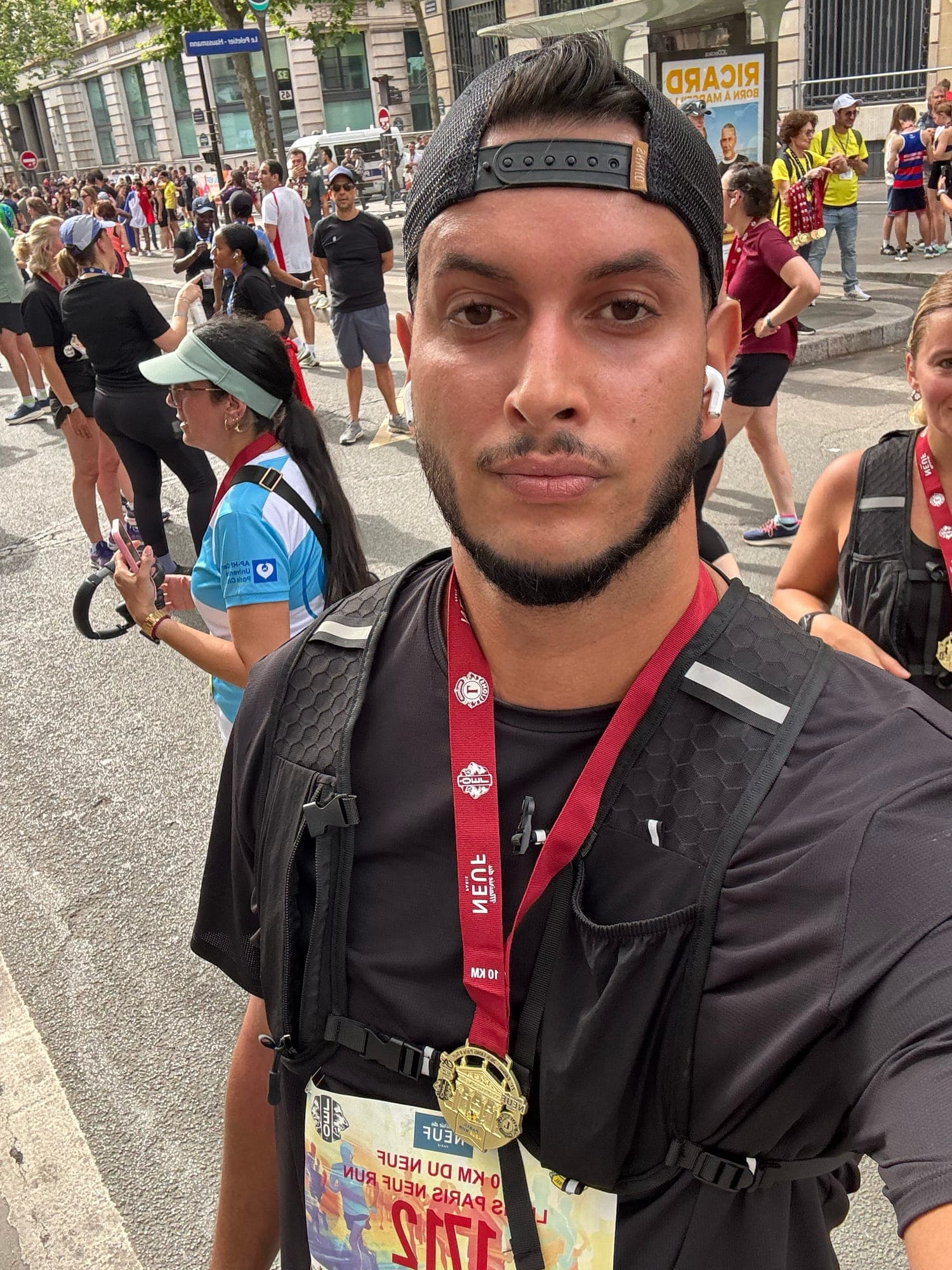 Selfie in a backwards cap and black running vest, with a gold medal on a red 10 km ribbon and bib 1712, runners in the street behind.