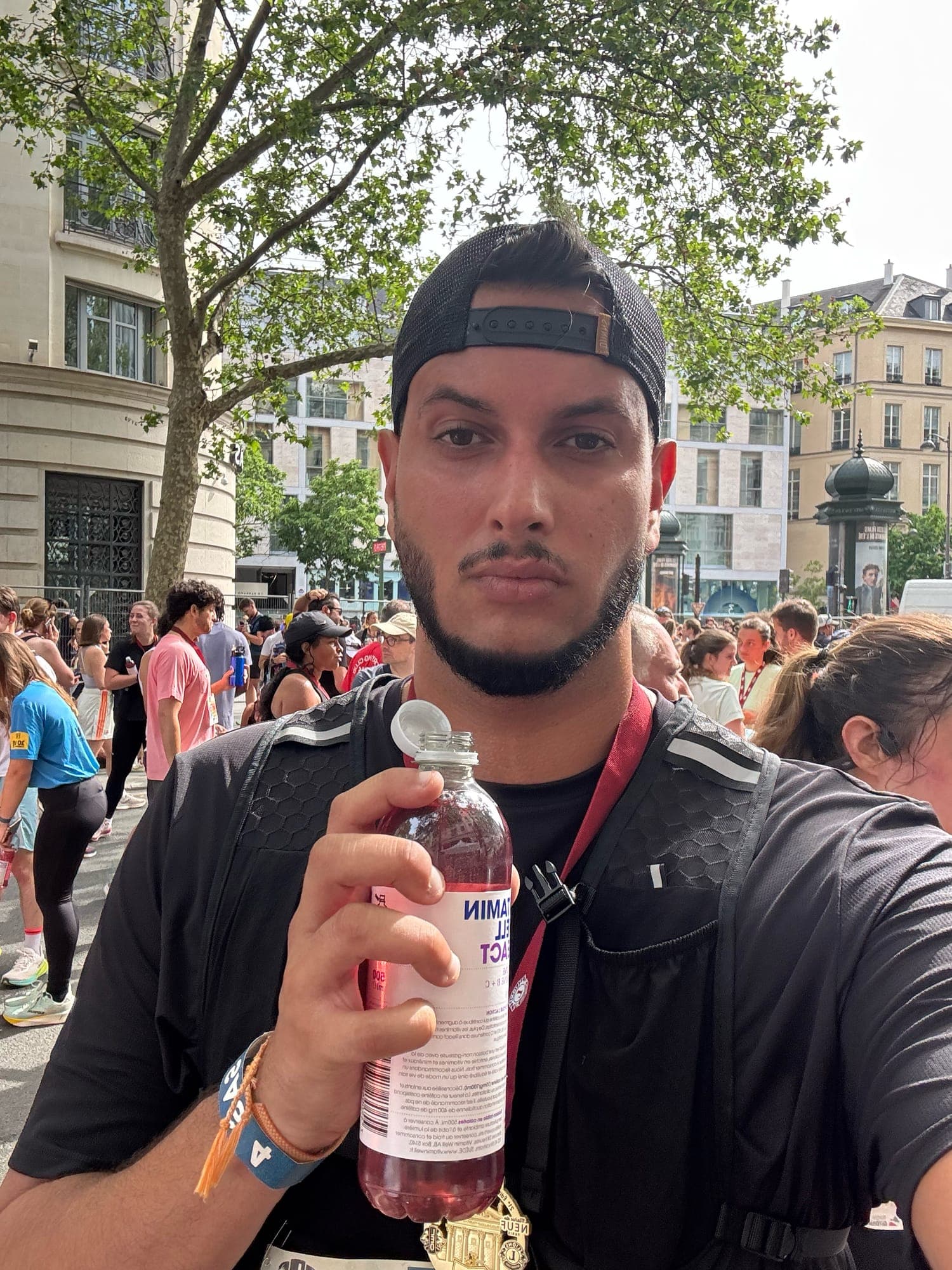 Selfie holding a pink drink bottle, a medal on a red ribbon around the neck, runners walking along a tree-lined street behind.