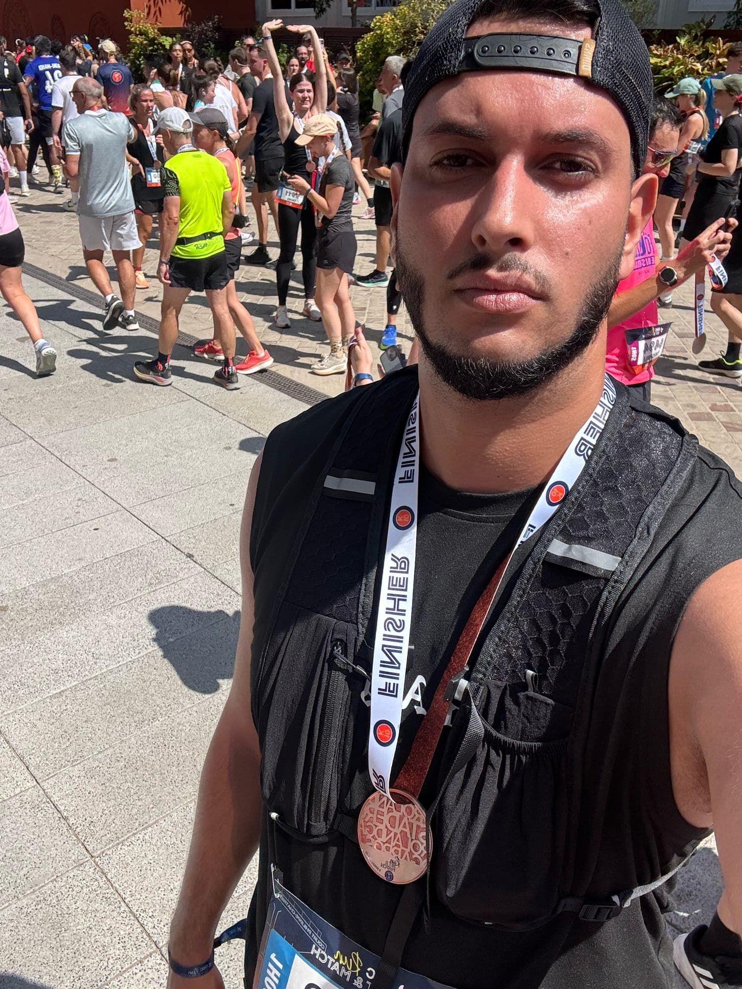 Selfie in a black running vest with a copper medal on a white Finisher lanyard, a crowd of runners behind.