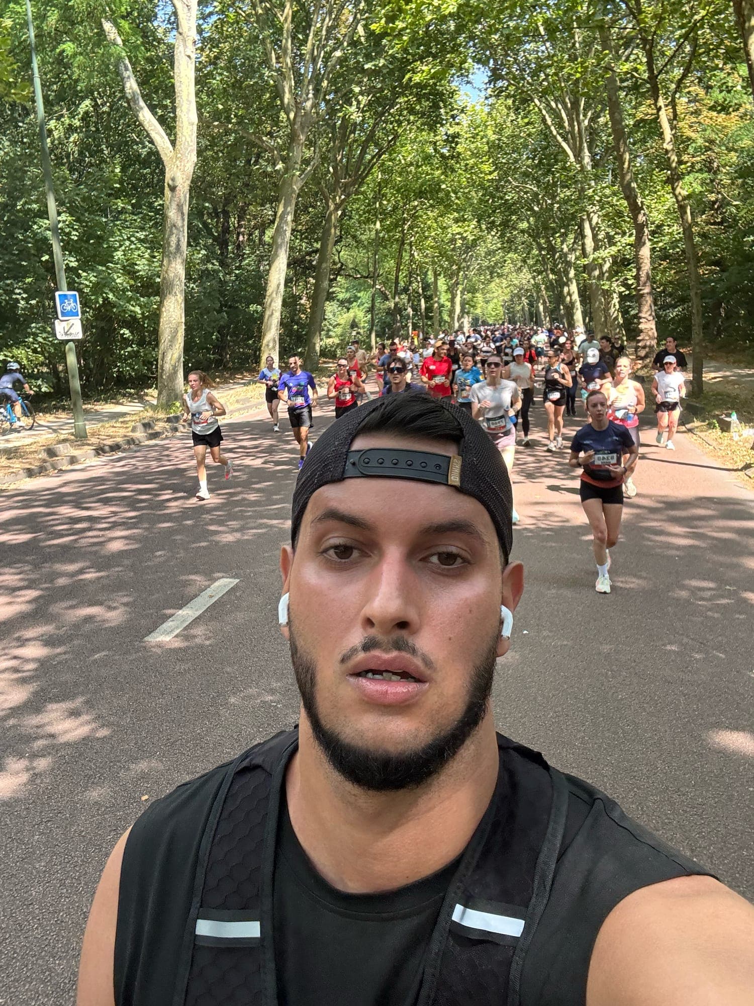 Selfie while running on a road through trees, a long line of runners wearing bibs behind.