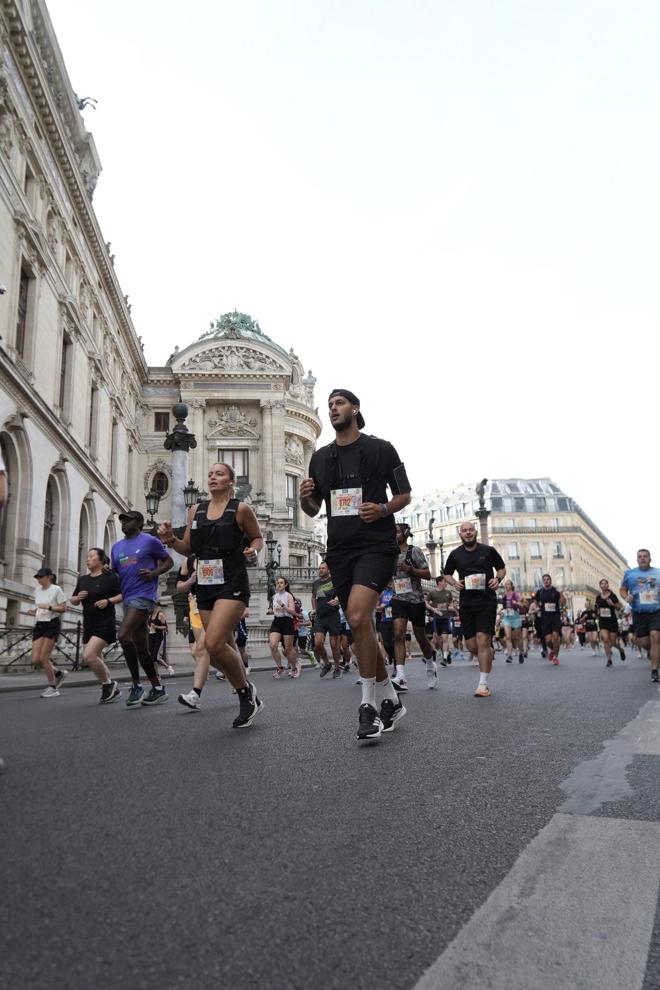 Runner in a black T-shirt, black running vest and backwards cap, bib 1712, running with other runners in front of the Paris opera house.